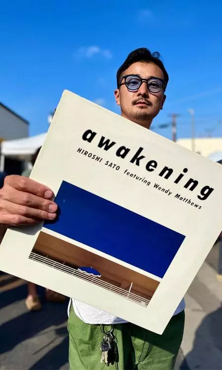 DJ Tsugu holding a copy of Hiroshi Sato&rsquo;s Awakening against a clear blue sky